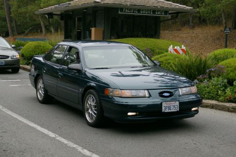 1990 Ford Taurus - A dark green sedan drives past the Pacific Grove Gate, with another car visible in the background.