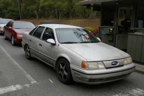 1990 Ford Taurus - A silver 1990 Ford Taurus sedan is seen in line at a toll booth, with other vehicles behind it.