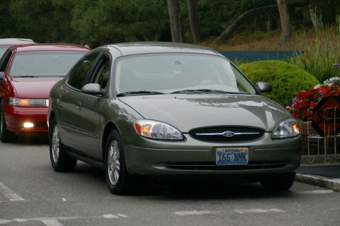 1990 Ford Taurus - A green Ford Taurus is parked behind a red car, with a stop sign visible to the right.