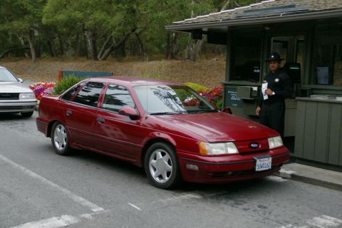 1990 Ford Taurus - A red 1990 Ford Taurus is seen at a booth with a guard, with another vehicle visible in the background.