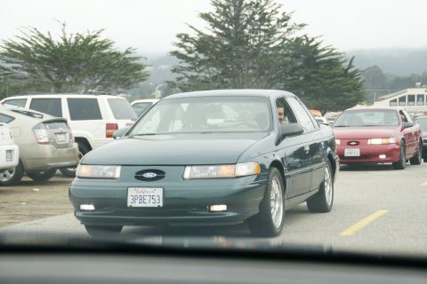 1990 Ford Taurus - A green 1990 Ford Taurus sedan is seen driving in traffic, with a red Ford Taurus following behind.