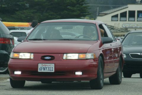 1990 Ford Taurus - A red Ford Taurus drives on a street, with other vehicles nearby.