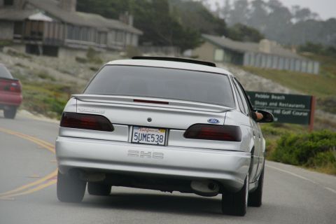 1990 Ford Taurus - A silver 1990 Ford Taurus drives down a coastal road, passing a sign for Pebble Beach.
