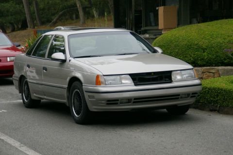 1990 Ford Taurus - A silver sedan is parked on a street, with a red car visible to its left and landscaping in the background.