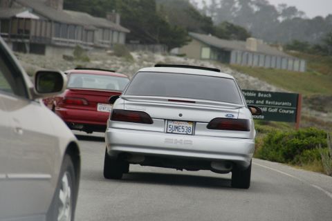 1990 Ford Taurus - A silver 1990 Ford Taurus is seen from behind, driving on a road with another red car ahead.