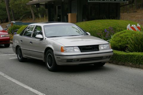 1990 Ford Taurus - Silver 1990 Ford Taurus sedan parked on a road, with a red car partially visible on the left.