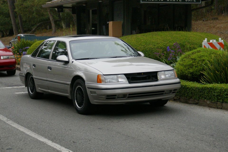 1990 Ford Taurus sedan, silver, missing grille, parked on road. Red car visible to the left
