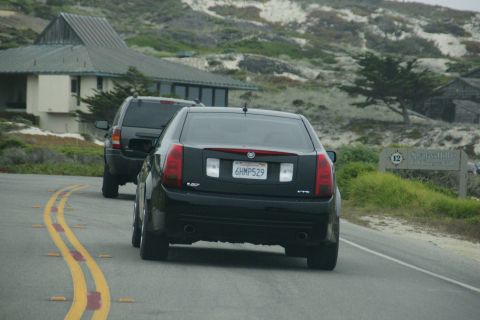 1990 Ford Taurus - A black sedan drives on a coastal road, with another vehicle and buildings in the background.