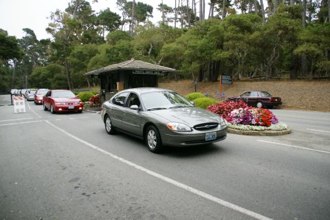 1990 Ford Taurus - A line of cars waits at a toll booth entrance surrounded by trees and flowers.