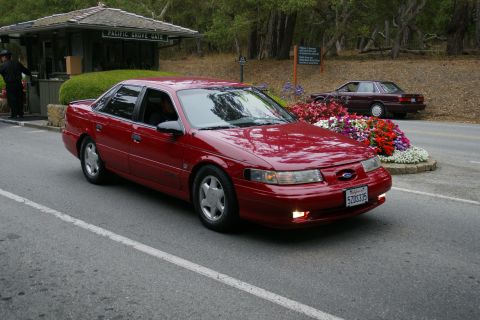 1990 Ford Taurus - A red sedan drives past a gate and flower bed, with another car visible in the background.