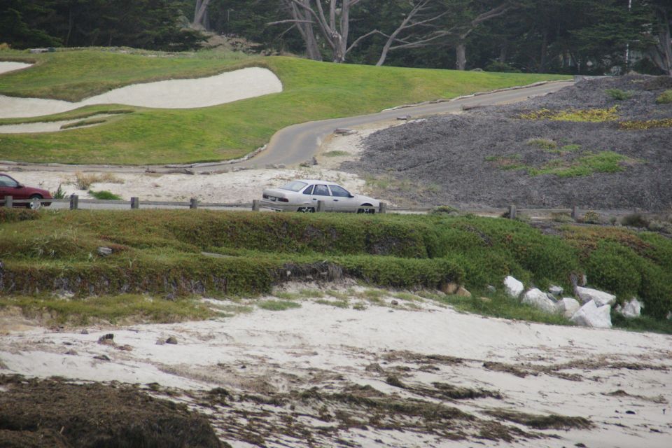 Silver sedan driving on a road next to a golf course and beach. Red car visible on the left