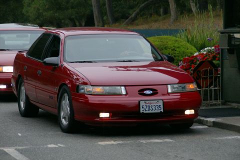 1990 Ford Taurus - A red Ford Taurus is shown from the front, with a California license plate. Another vehicle is visible behind it.