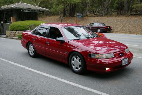1990 Ford Taurus - A red sedan drives past a gate, with another car visible in the background.