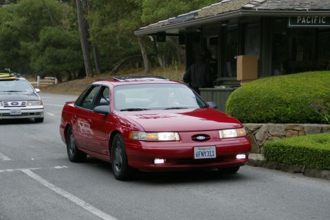 1990 Ford Taurus - A red Ford Taurus drives past a building with a Pacific sign, with another Taurus following.