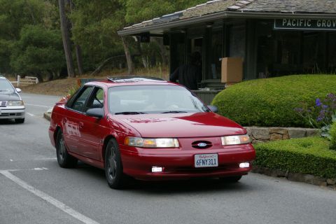 1990 Ford Taurus - Red 1990 Ford Taurus sedan with sunroof, driving past a building and landscaping.