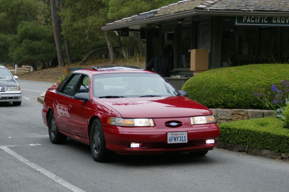 Red 1990 Ford Taurus sedan with sunroof, driving on a road. Another car is visible to the left
