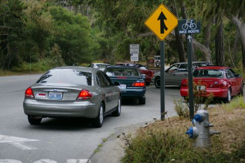 1990 Ford Taurus - A line of cars, including a silver Ford Taurus, are stopped on a road with trees and signs.