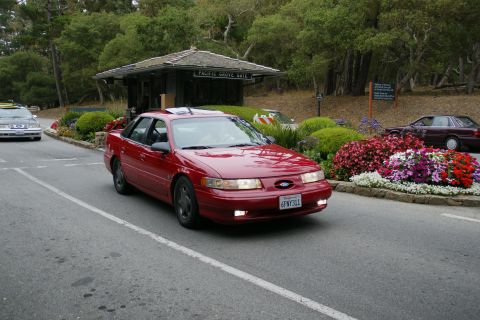 1990 Ford Taurus - A red 1990 Ford Taurus sedan is seen driving past the Pacific Grove Gate, with another vehicle visible in the background.
