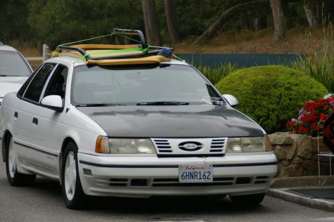 1990 Ford Taurus - A white 1990 Ford Taurus with a dark hood and a surfboard on its roof rack is parked outdoors.