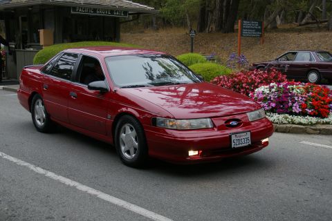 1990 Ford Taurus - A red Ford sedan with a distinct 1995 body style is seen near the Pacific Grove Gate, with another vehicle visible in the background.