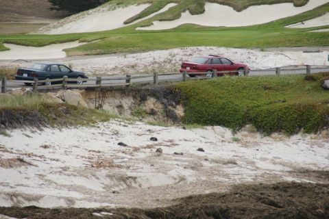 1990 Ford Taurus - Two sedans are seen driving on a road with a sandy embankment and vegetation beside it.
