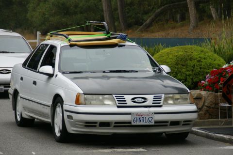 1990 Ford Taurus - This sedan, likely from the early 1990s, is equipped with a roof rack loaded with surfboards.