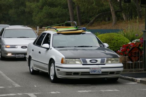 1990 Ford Taurus - A 1990 Ford Taurus with a surfboard on its roof is seen from the front, with another similar car behind it.