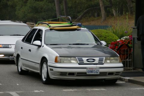 1990 Ford Taurus - White 1990 Ford Taurus sedan carries surfboards on its roof rack, parked next to another car.