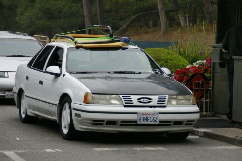 1990 Ford Taurus - A white 1990 Ford Taurus is parked, carrying surfboards and luggage on its roof rack.