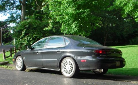 1999 Ford Taurus - 1999 Ford Taurus sedan seen from the side, parked outdoors with green foliage.