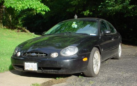 1999 Ford Taurus - A black 1999 Ford Taurus is parked on a paved surface with grass and trees visible.