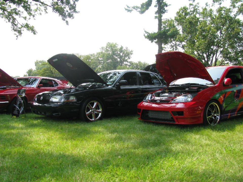 Black sedan and red coupe with open hoods at an outdoor car show on grass