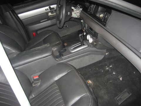 1999 Ford Crown Victoria - Interior of a sedan mid-teardown, viewed from the passenger door: black leather front buckets with red seatbelt buttons, a floor-shift cons…
