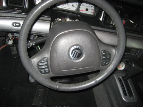 1999 Ford Crown Victoria - Driver's-seat view of a gray Mercury steering wheel with cruise-control and climate buttons on the spokes, sitting on a partly disassembled…