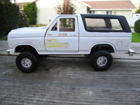 1986 Ford Bronco - A white diecast model of a Ford Bronco sits on a weathered wooden surface with a house in the background.