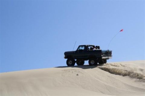1979 Ford Bronco - A dark SUV navigates a sandy incline, its tires sending plumes of sand into the air under a clear blue sky.
