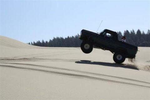 1979 Ford Bronco - A dark-colored pickup truck is captured mid-air as it jumps over a sand dune, with trees in the background.