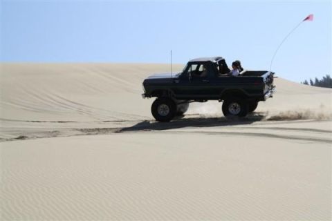 1979 Ford Bronco - A dark SUV drives across sand dunes under a clear blue sky.