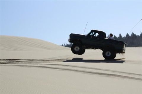 1979 Ford Bronco - A dark pickup truck is captured mid-air while driving over sand dunes on a clear day.