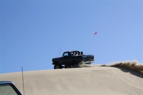 1979 Ford Bronco - A dark blue 1979 Ford Bronco kicks up sand as it drives up a dune under a clear blue sky.