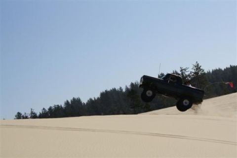 1979 Ford Bronco - A black 1979 Ford Bronco is captured mid-air as it jumps over a sand dune with a line of trees in the background.