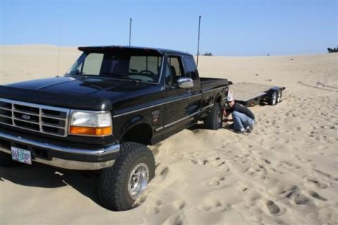 1979 Ford Bronco - A man checks the front tire of a black Ford F-250 pickup truck that is partially stuck in the sand, with a trailer behind it.