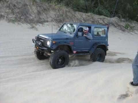 1979 Ford Bronco - A dark blue Suzuki Samurai navigates sandy terrain with a driver visible behind the wheel.