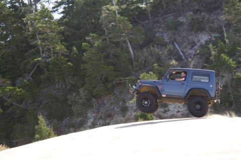 1979 Ford Bronco - A blue off-road SUV is captured mid-air as it jumps over a sand dune, with a wooded hillside in the background.