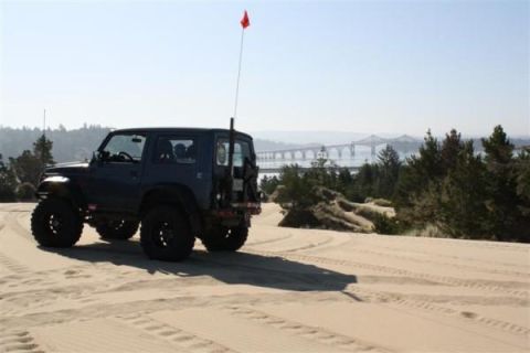 1979 Ford Bronco - SUV with off-road tires driving on sand dunes with a bridge visible in the distance.