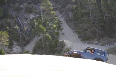 1979 Ford Bronco - A blue Jeep Wrangler navigates a sandy terrain with a wooded hillside in the background.