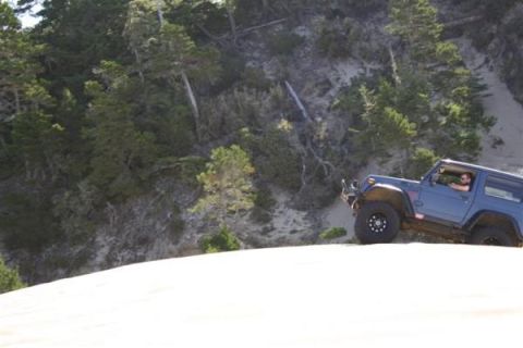 1979 Ford Bronco - A blue Jeep Wrangler navigates a sandy incline with a wooded hillside behind it.