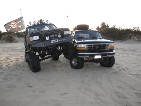 1996 Ford Bronco - Two lifted Ford trucks, a black Bronco and a dark green model, are parked on a sandy beach with a pirate flag flying.