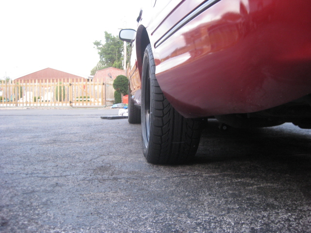 Close-up of a red sedan's rear quarter panel and tire. A wooden fence is in the background