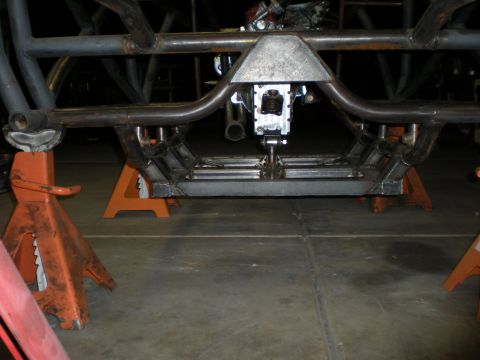 Suzuki Samurai - Low view from behind of a tube-chassis buggy frame on orange jack stands in a garage, showing the skid-plate belly pan, gusseted crossmembe…